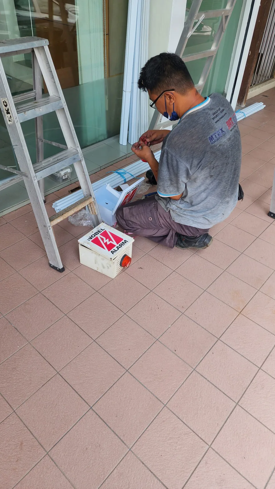 Technician assembles an alarm component beside a ladder and equipment box.