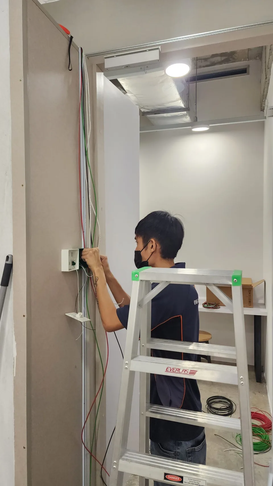 Technician on a ladder connecting a wall reader and overhead wiring.