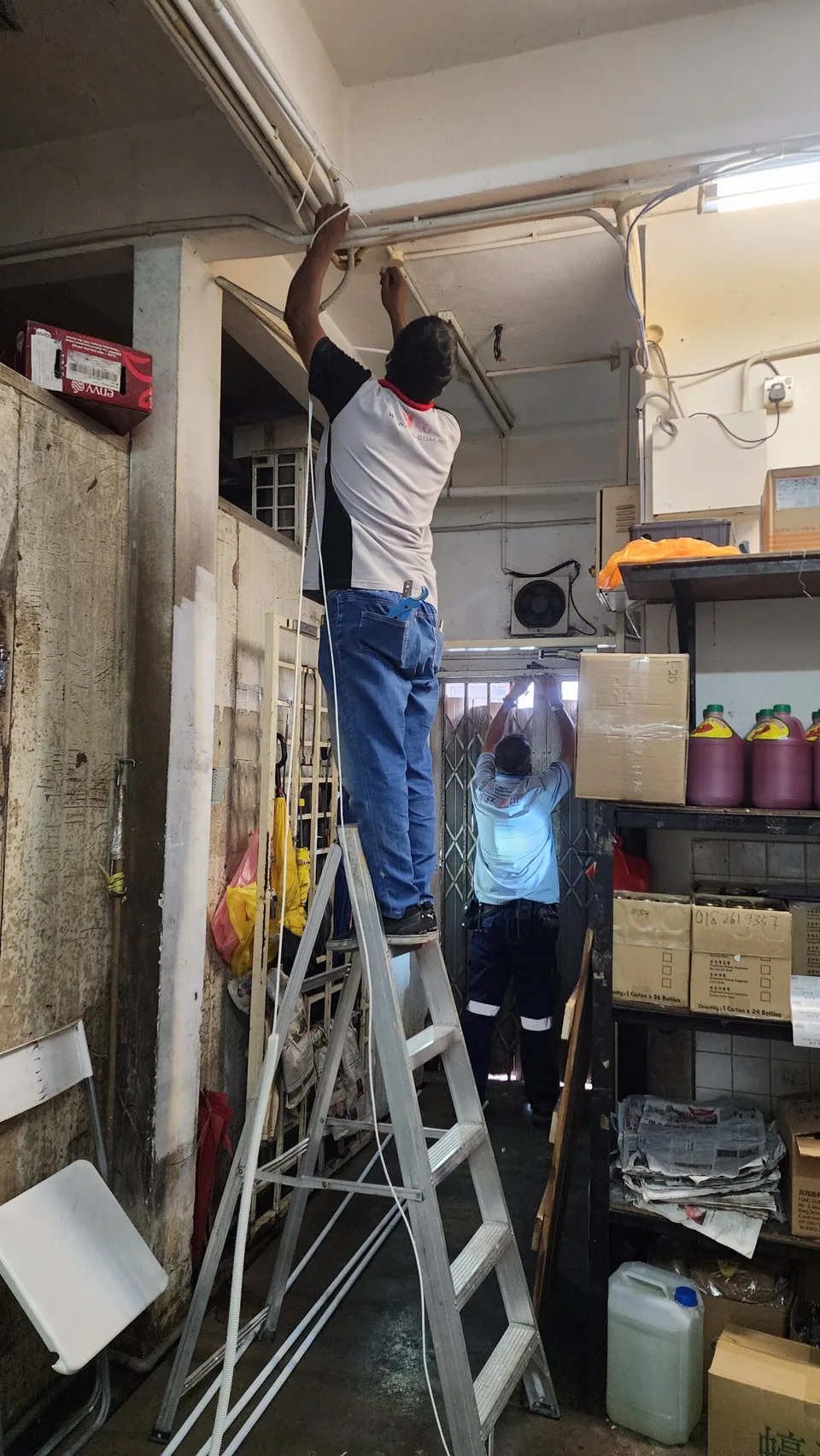 Two technicians route conduit and wiring across a storeroom ceiling.