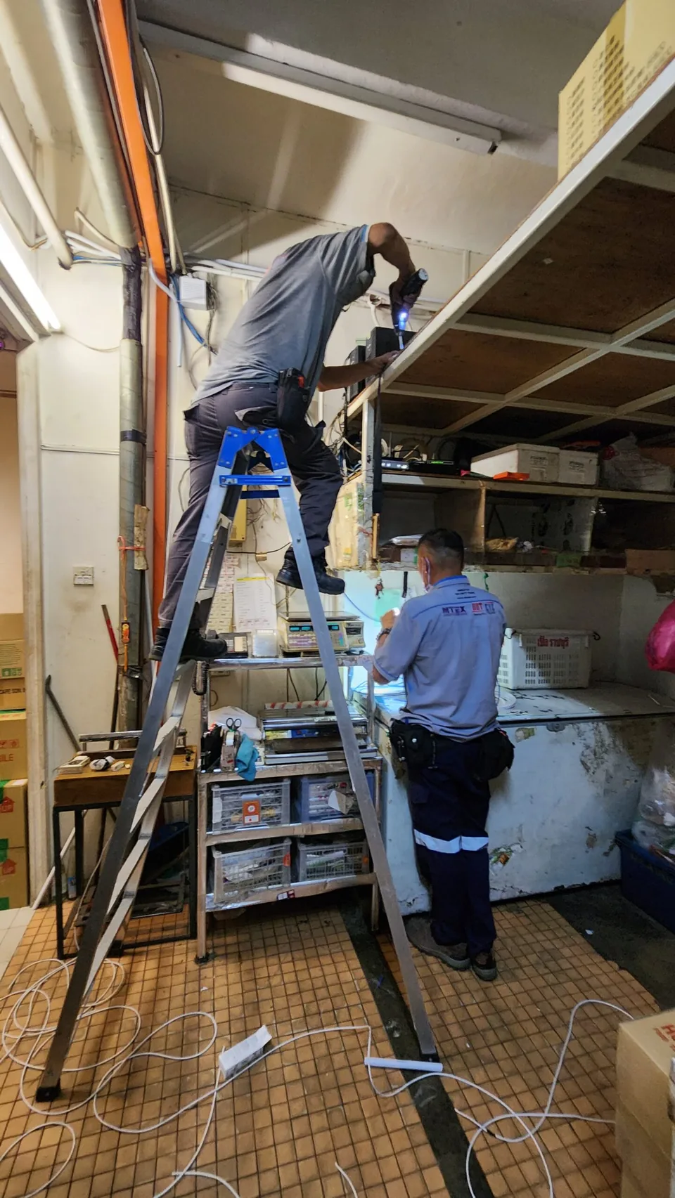 Two technicians work with cables and a ladder in a storeroom.